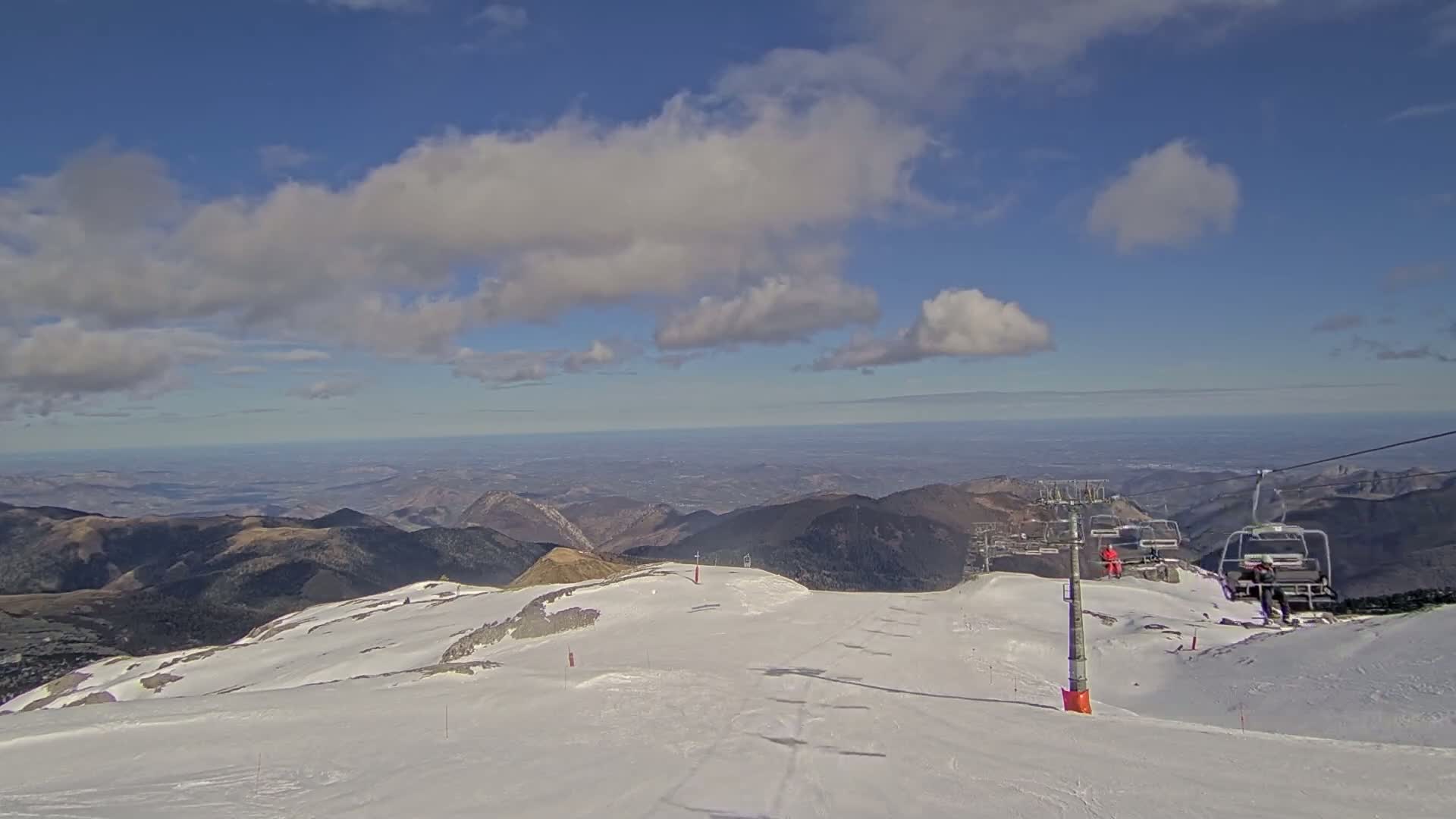 A snowy mountaintop ski slope with a chairlift and distant valley views under a partly cloudy blue sky.
