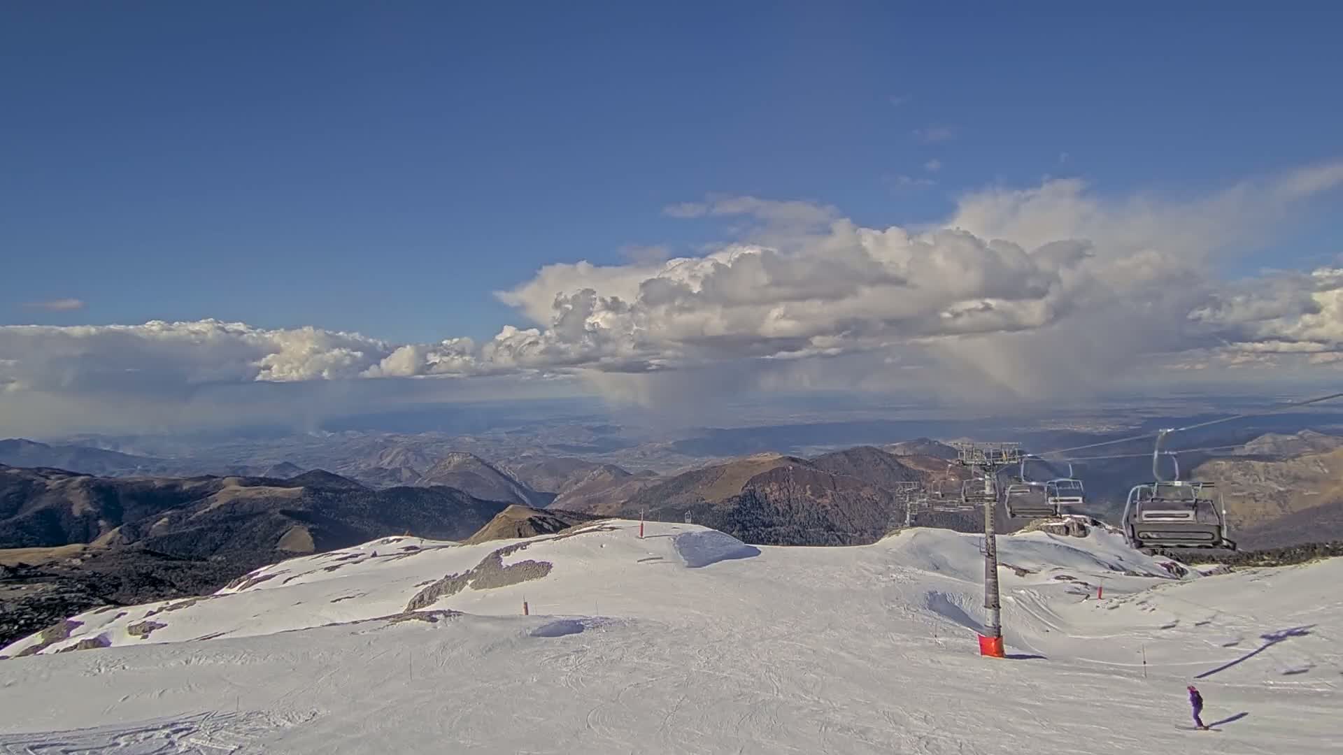 A snow-covered mountaintop ski slope with a chairlift and a skier, under a partly cloudy sky with rain showers in the distance.