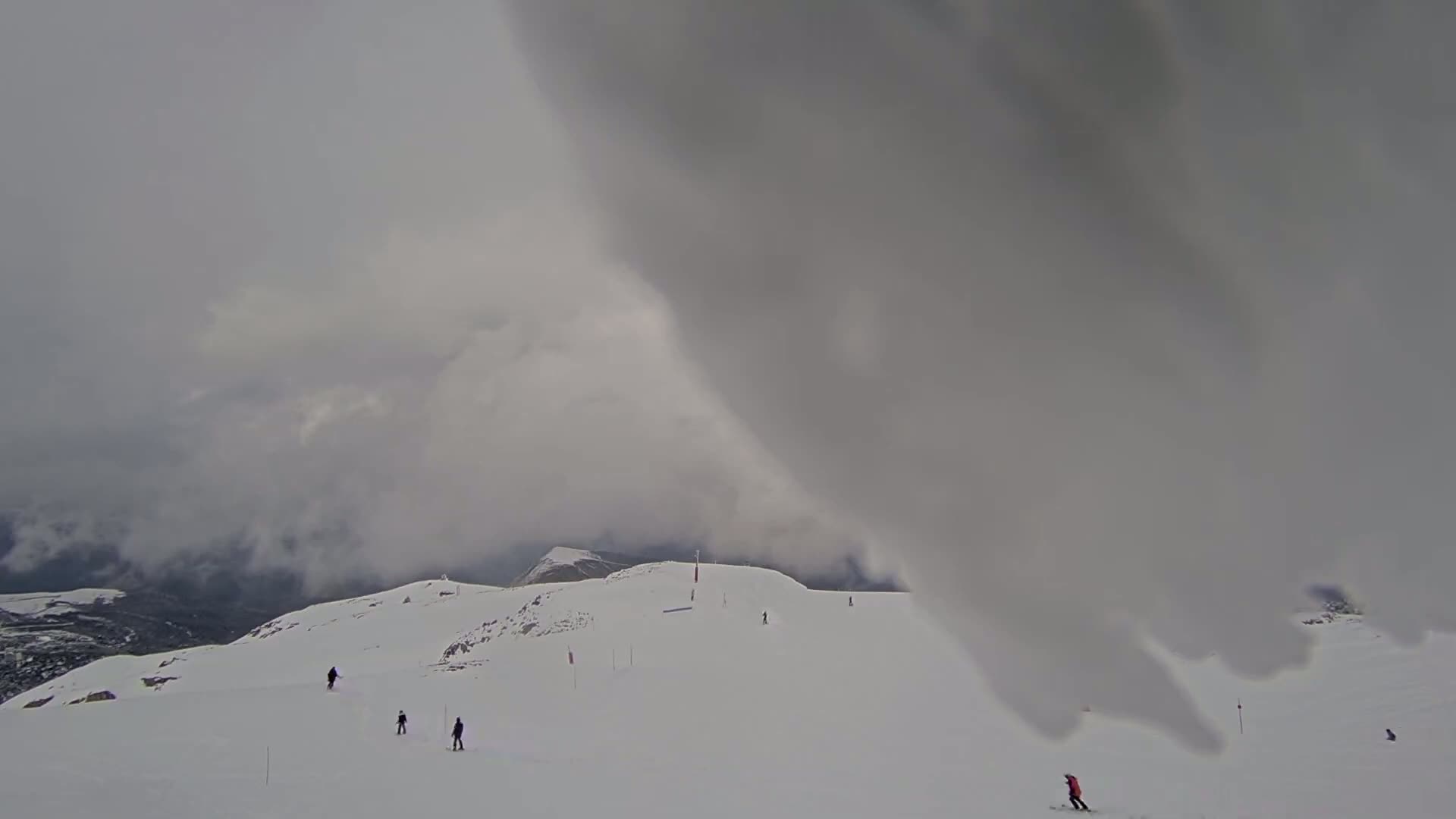 A snowy mountain slope with several skiers visible under a cloudy, overcast sky.