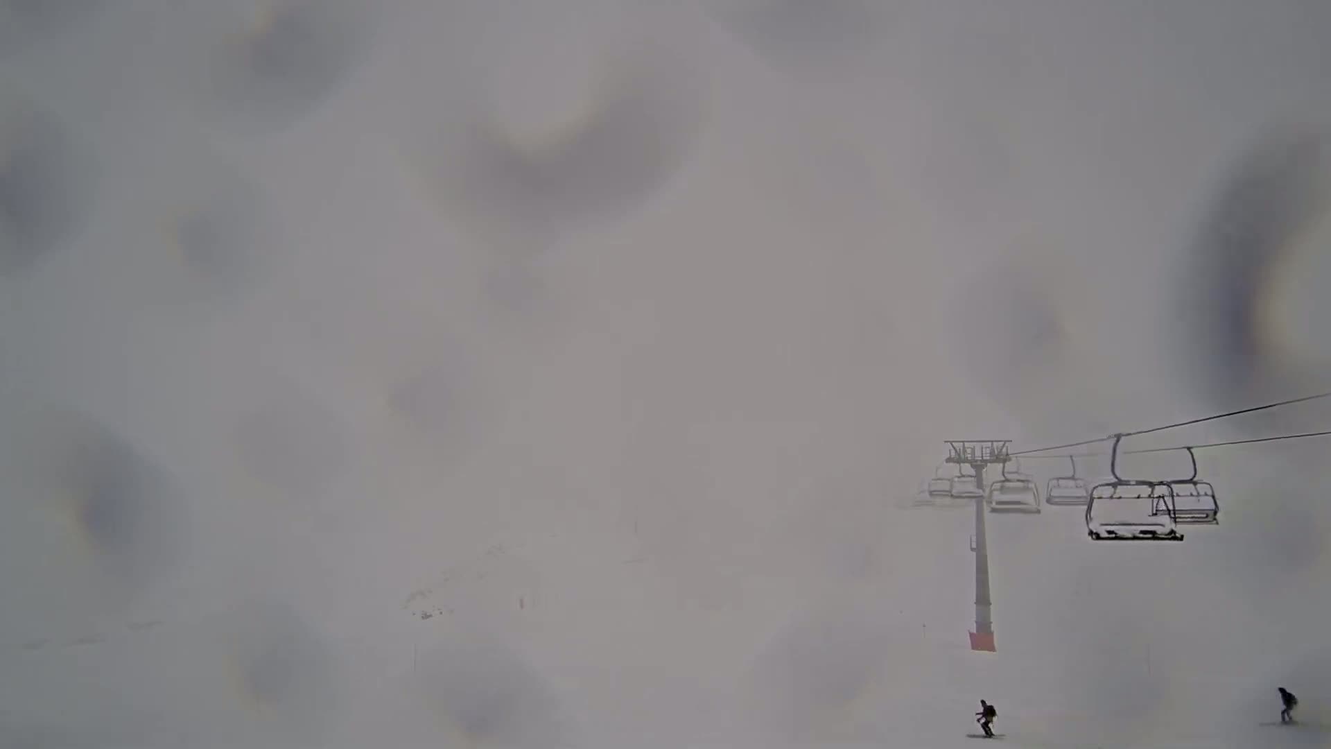 A ski lift with several empty chairs ascends through a dense, snowy fog, with two skiers visible in the distance.
