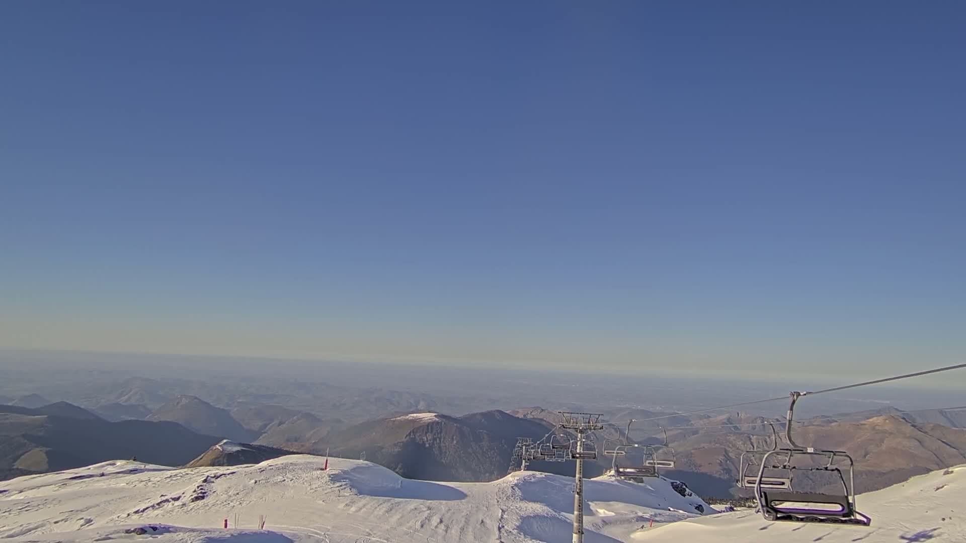 A chairlift ascends a snow-covered mountain range under a clear blue sky, overlooking a vast, hazy valley.