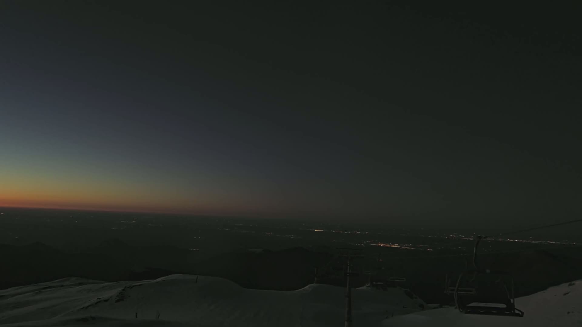 A snow-covered mountaintop at twilight overlooks a distant city illuminated by lights under a mostly clear, dark sky.