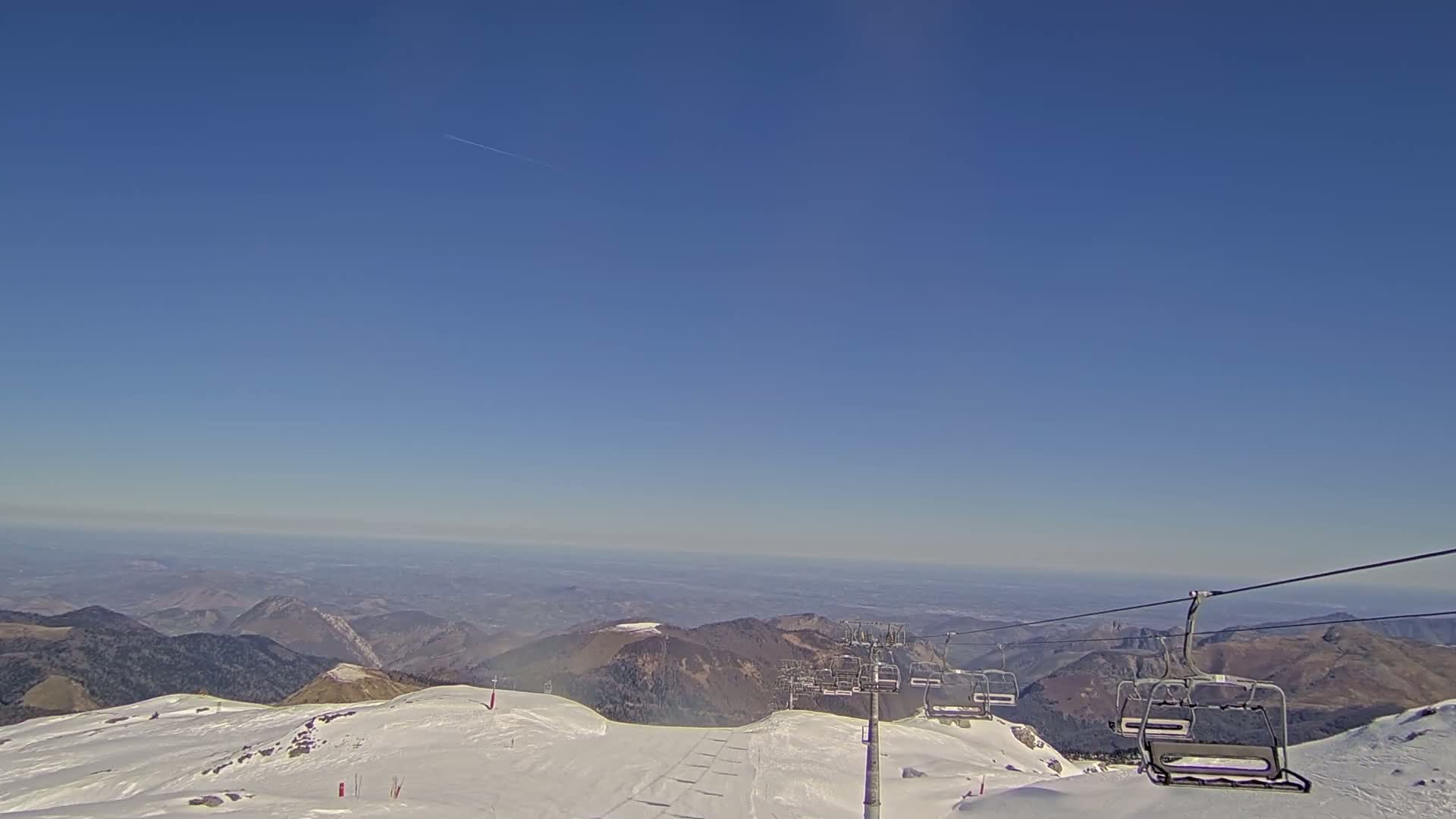 A ski lift ascends a snow-covered mountain range on a clear, sunny day, overlooking a vast valley.