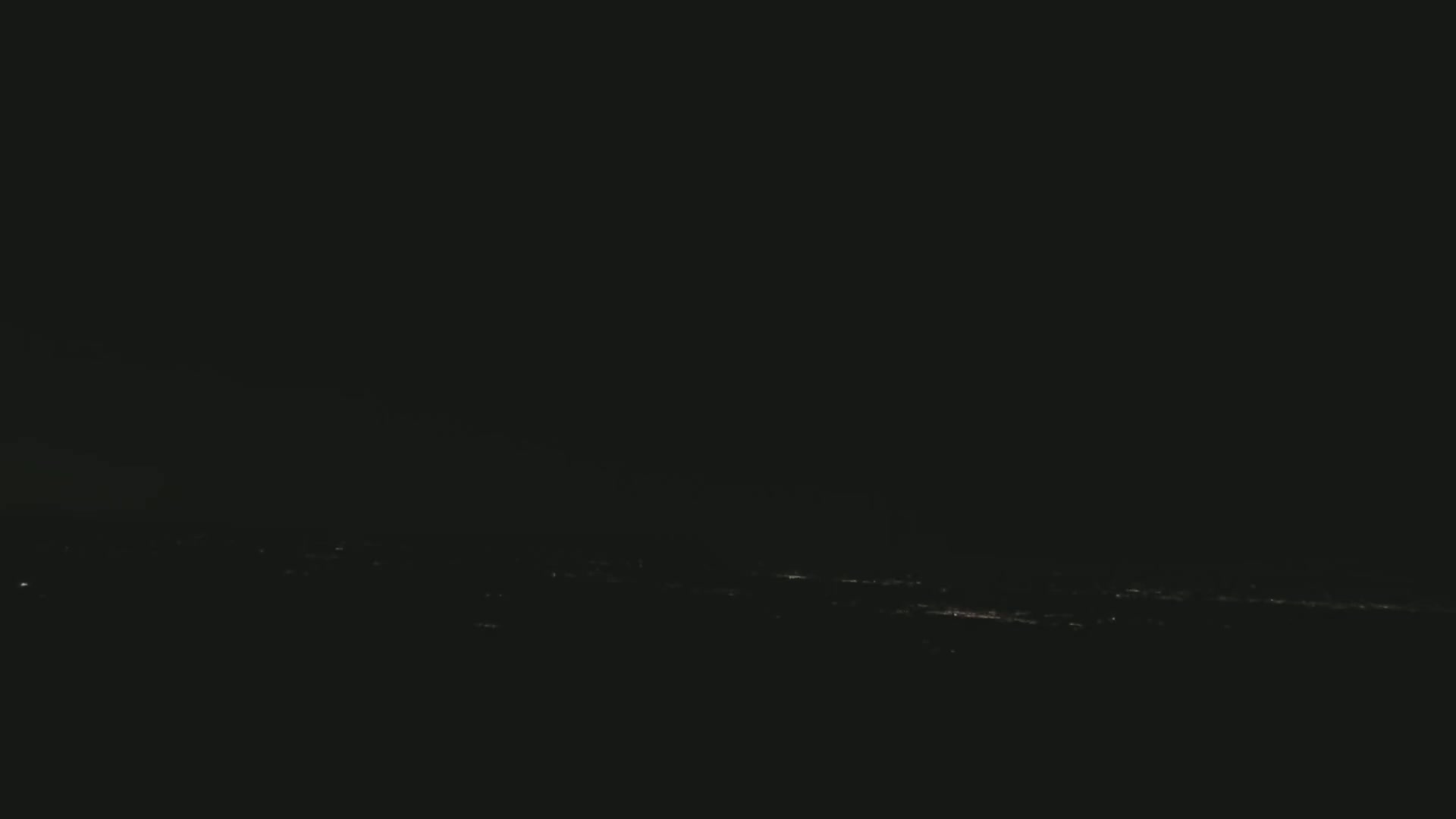 A dark nighttime scene shows a distant, faintly lit cityscape under a mostly clear sky.