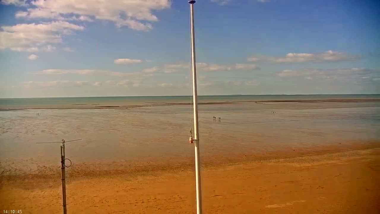A mostly sunny day shows a wide expanse of flat, wet sand extending to a calm ocean under a partly cloudy sky, with a tall flagpole and a smaller antenna in the foreground.