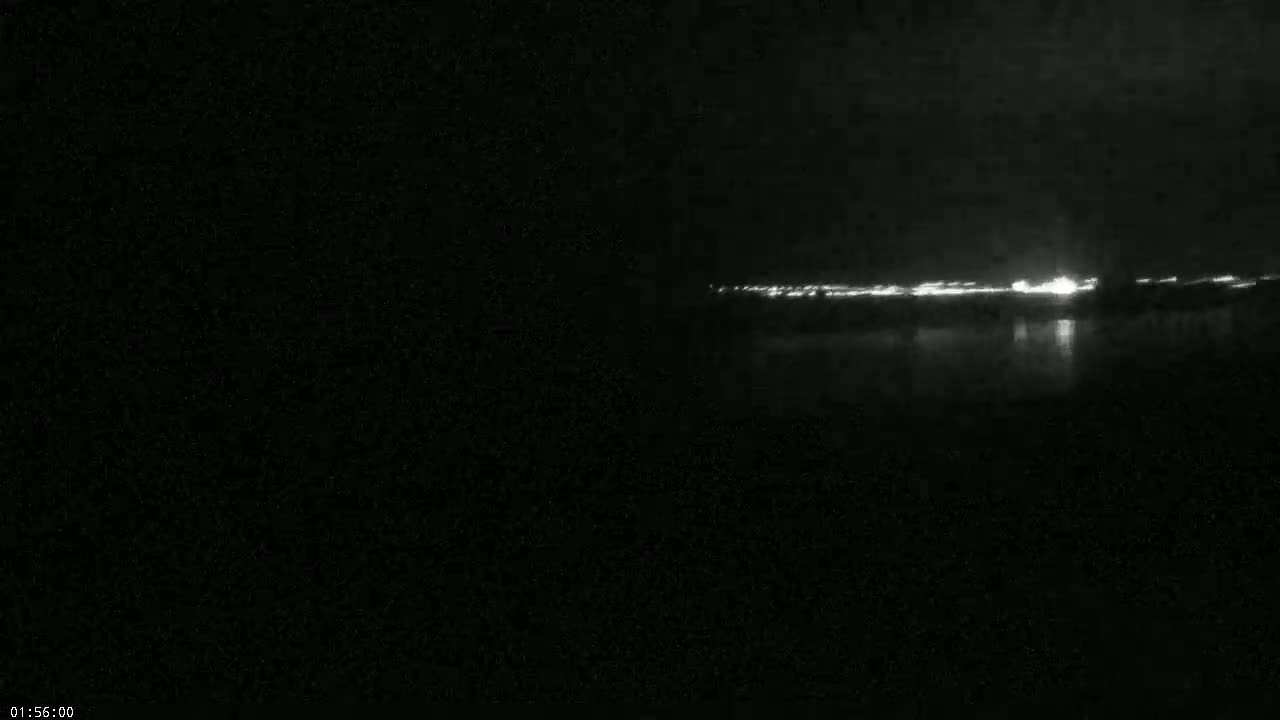 A dark, nighttime scene shows a distant line of lights reflecting on a body of water.