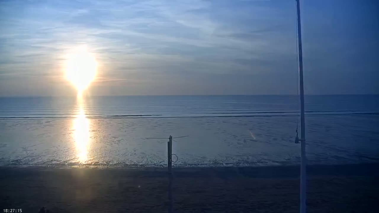 A setting sun casts a bright reflection on a calm ocean at a beach, with a tall pole and a shorter antenna visible in the foreground.