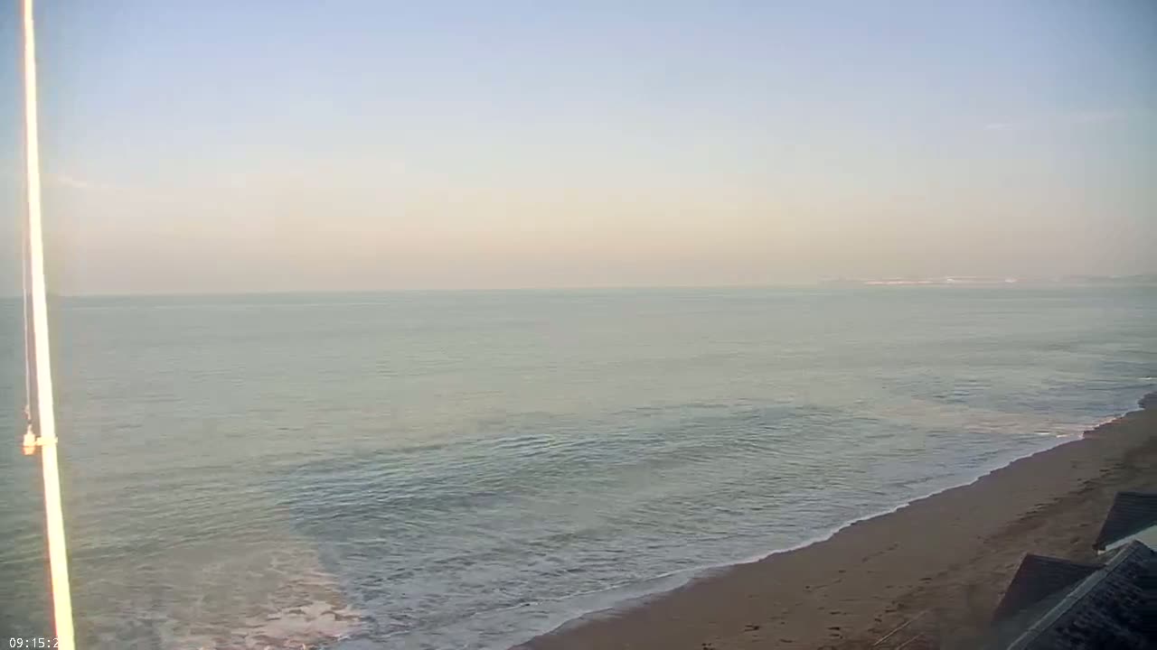 A sandy beach meets a calm, light-grey sea under a clear, pale sky.