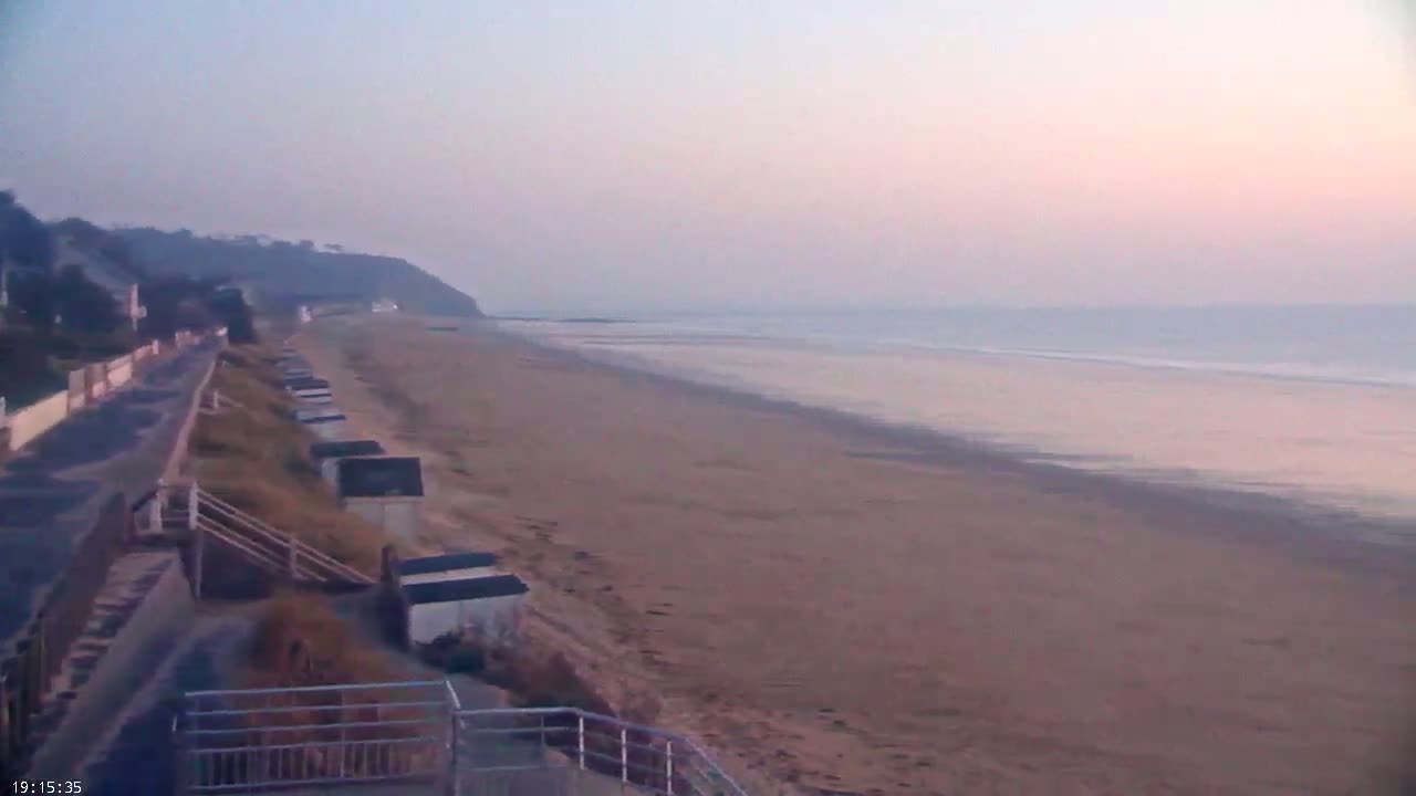 A long, sandy beach stretches out to a calm, hazy ocean under a pale pink and purple sunrise.