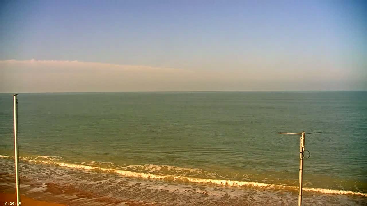 A mostly clear sky overlooks a calm sea with small waves lapping a sandy beach, and two tall poles stand on the shore.
