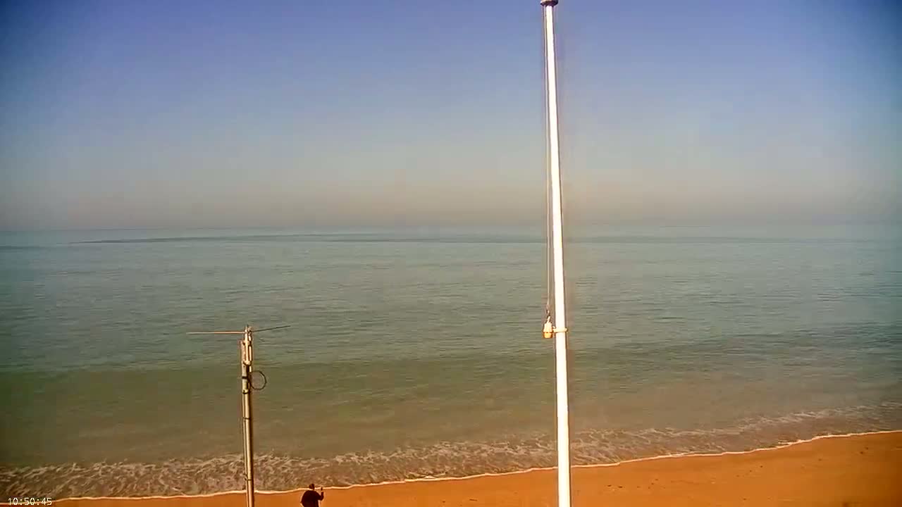 A calm sea with gentle waves laps a sandy beach under a clear, bright sky; a person stands on the beach near two tall poles.