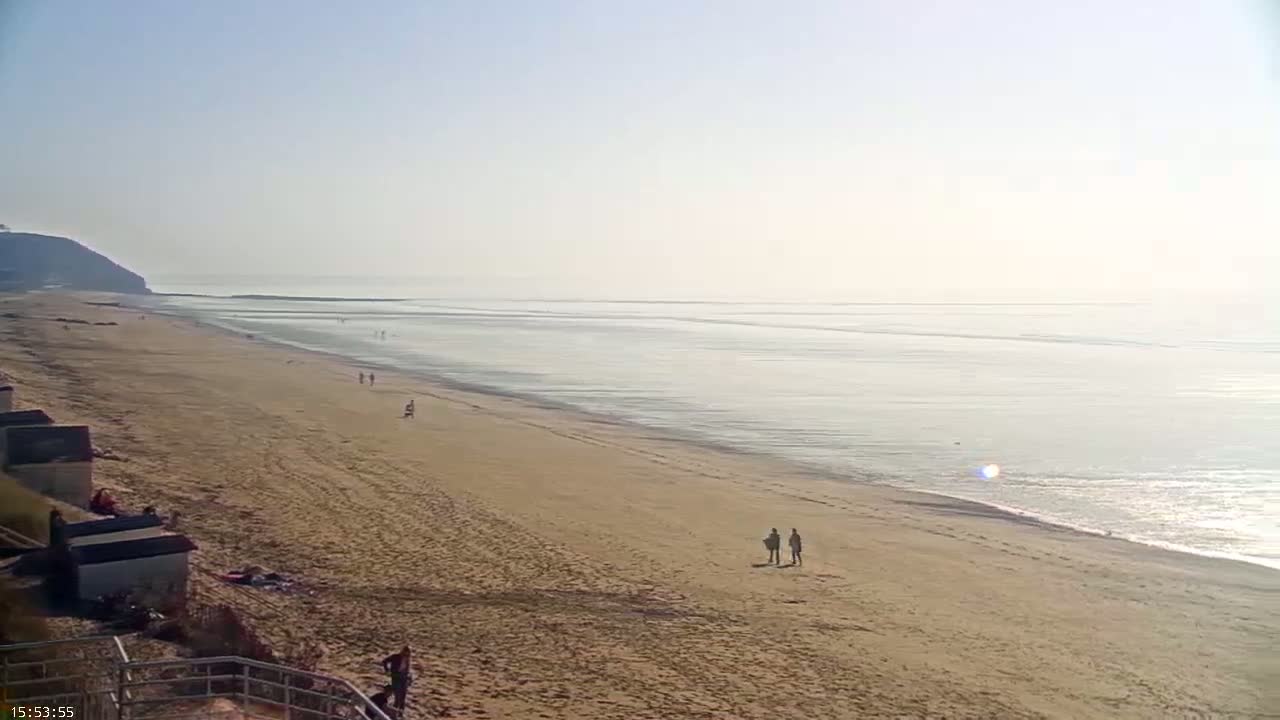 A long, sandy beach stretches towards a calm, light-grey sea under a clear, bright sky.