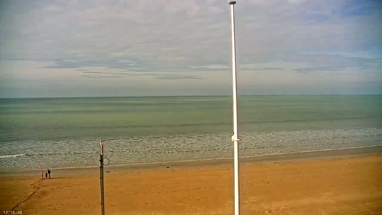 A sandy beach stretches towards a calm, greenish-blue sea under a mostly cloudy sky, with three people walking near the water's edge.