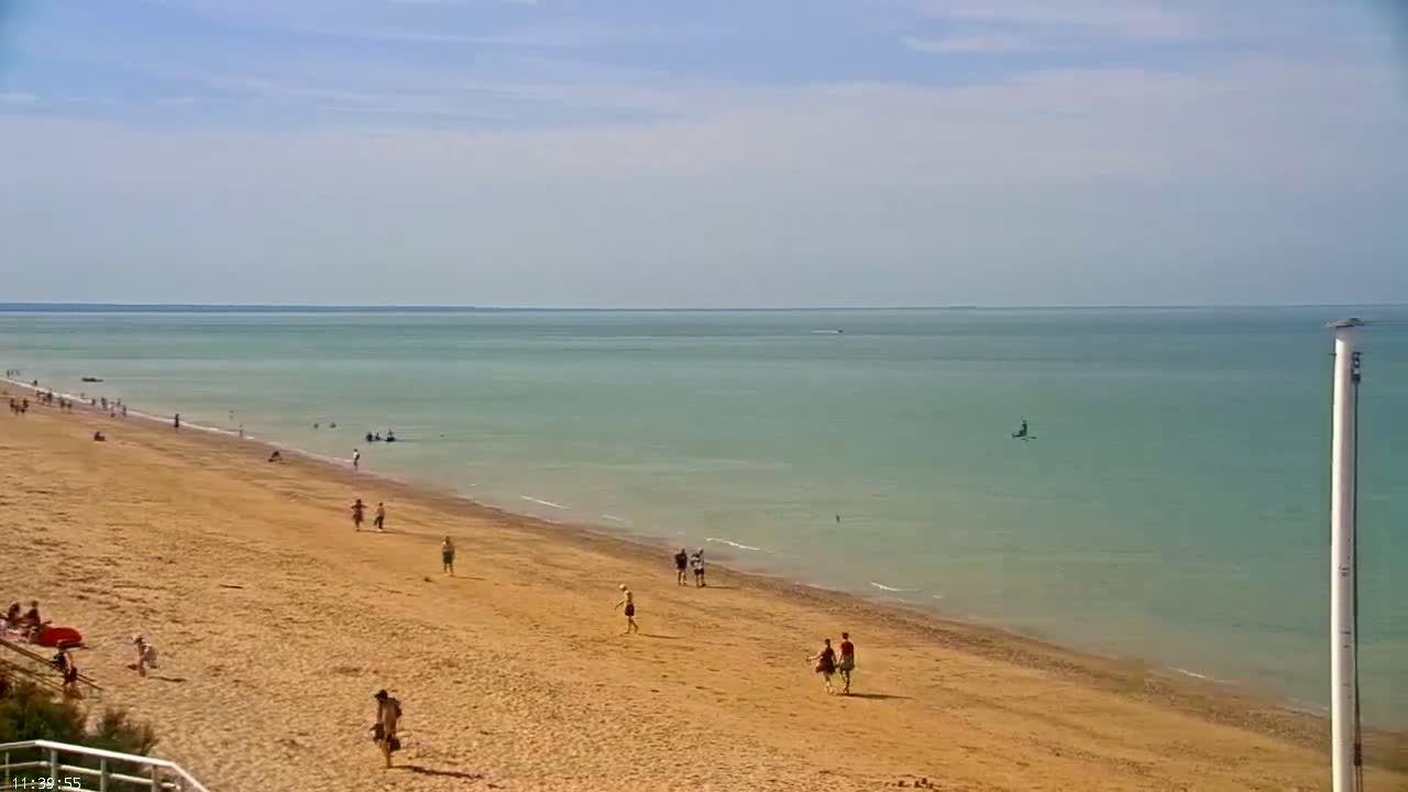 A sandy beach with a few people on it slopes down to a calm, light-green ocean under a bright, sunny sky.