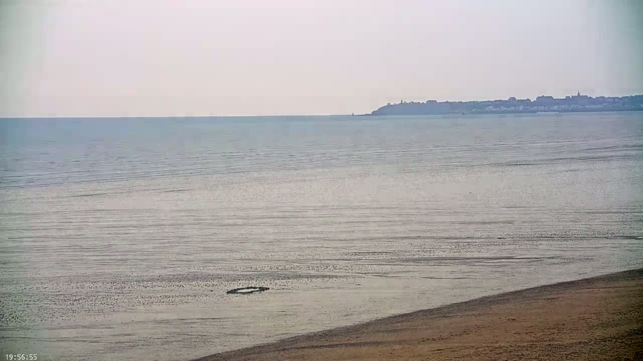 A sandy beach slopes down to a calm, light-grey sea, with a distant town visible on the horizon under a hazy sky.