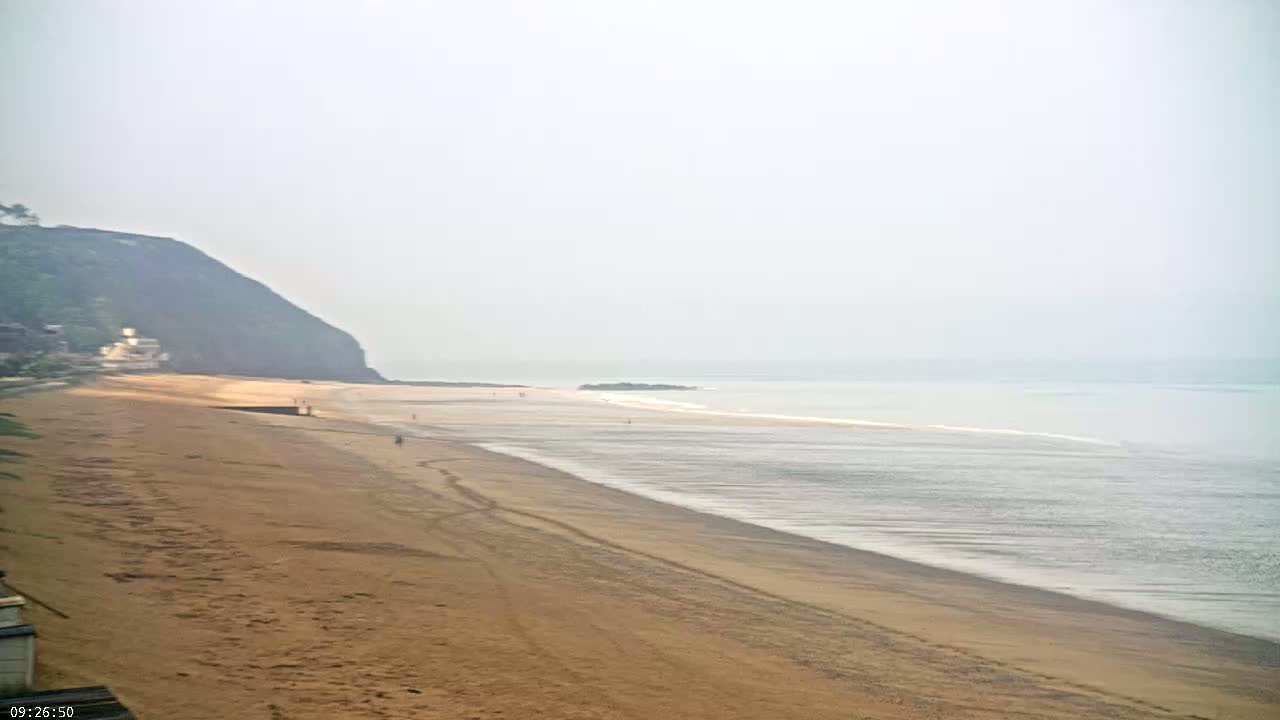 A hazy, light-colored beach stretches out to a calm sea, backed by a verdant cliff.