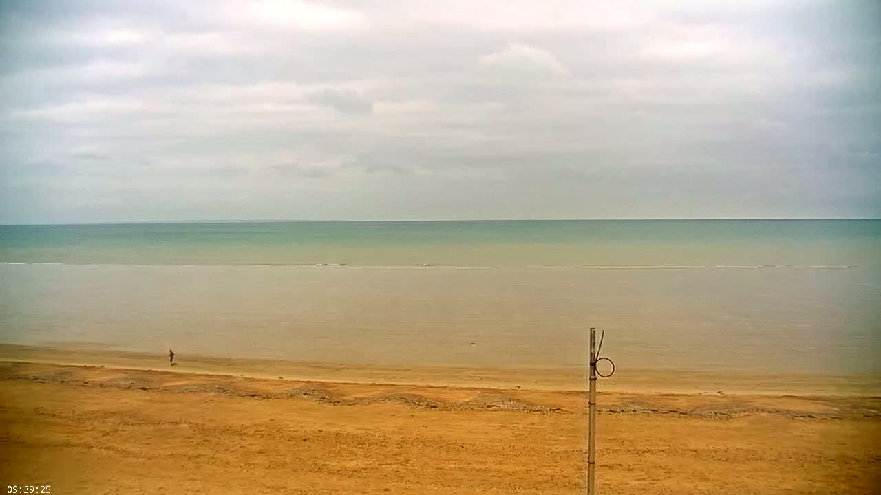 A person walks on a wide, sandy beach overlooking a calm, pale green-blue sea under a cloudy sky.