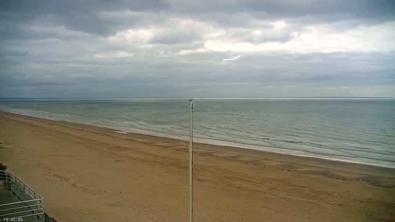 A long, sandy beach stretches towards a calm, grey sea under a cloudy sky.