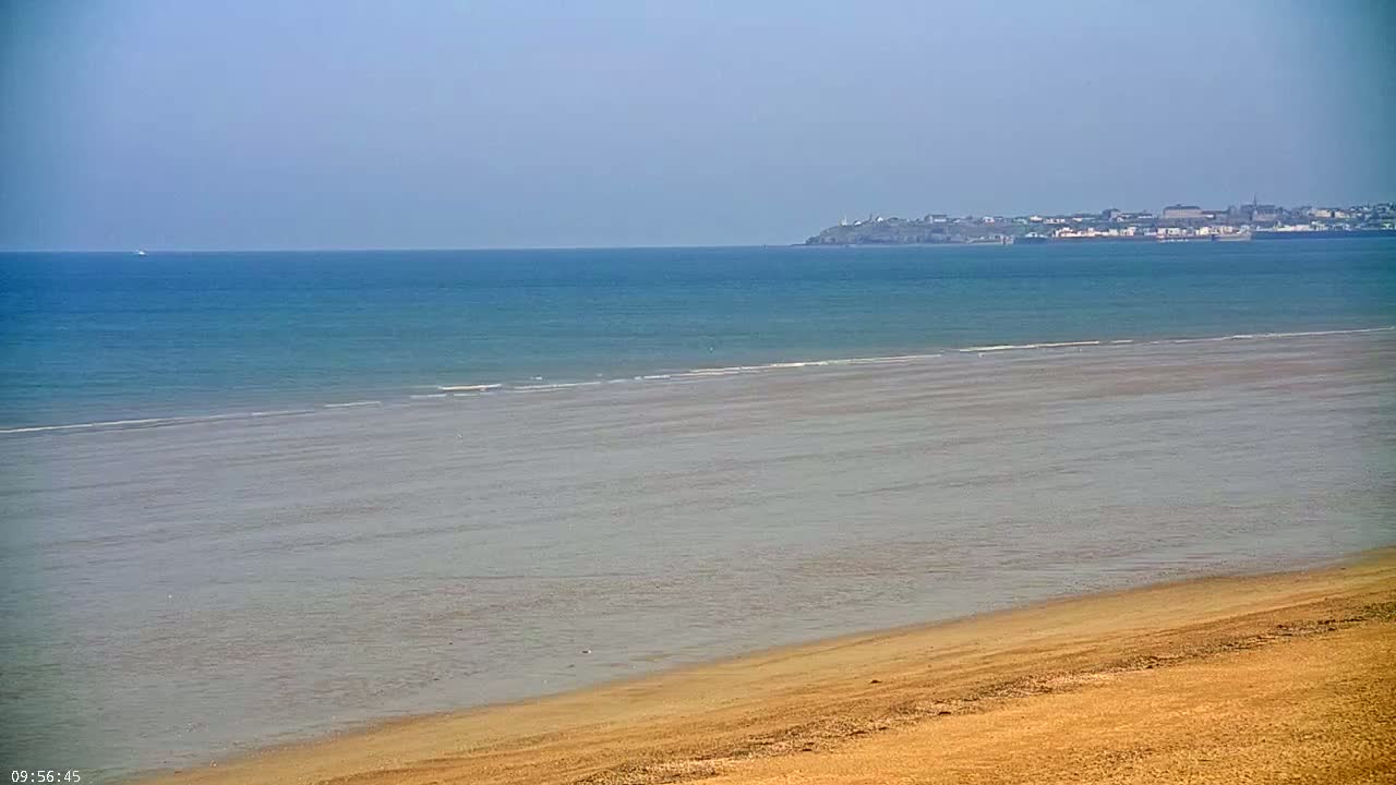 A sandy beach slopes down to a calm, teal ocean meeting a distant town under a clear sky.