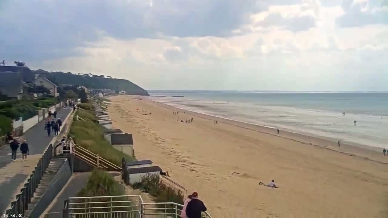 A long sandy beach stretches out to a calm ocean under a partly cloudy sky, with people walking on a path above the beach and others scattered on the sand.