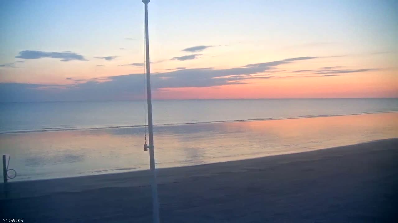 A calm ocean beach at sunset displays a pink and orange sky reflected in the water.