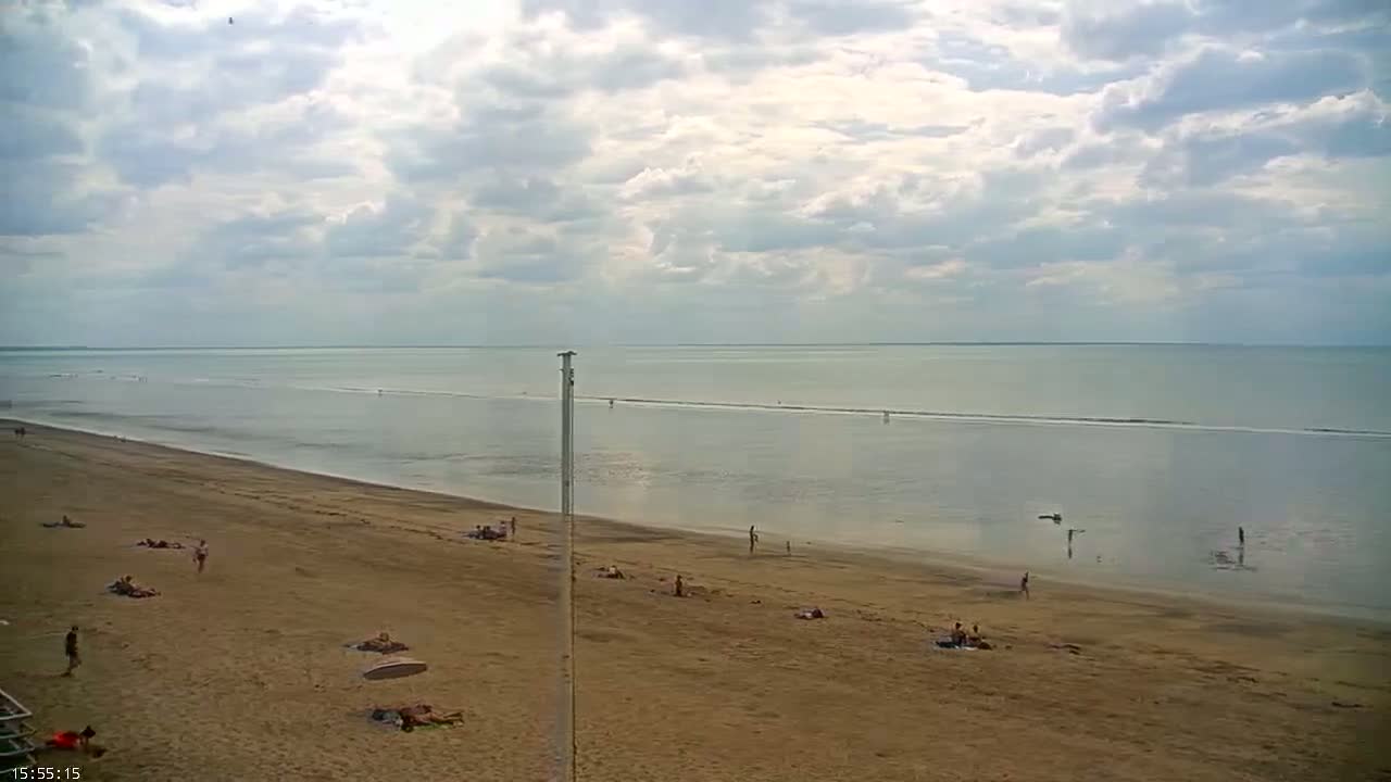 A wide sandy beach with scattered people sunbathing and walking, extends to a calm sea under a partly cloudy sky.