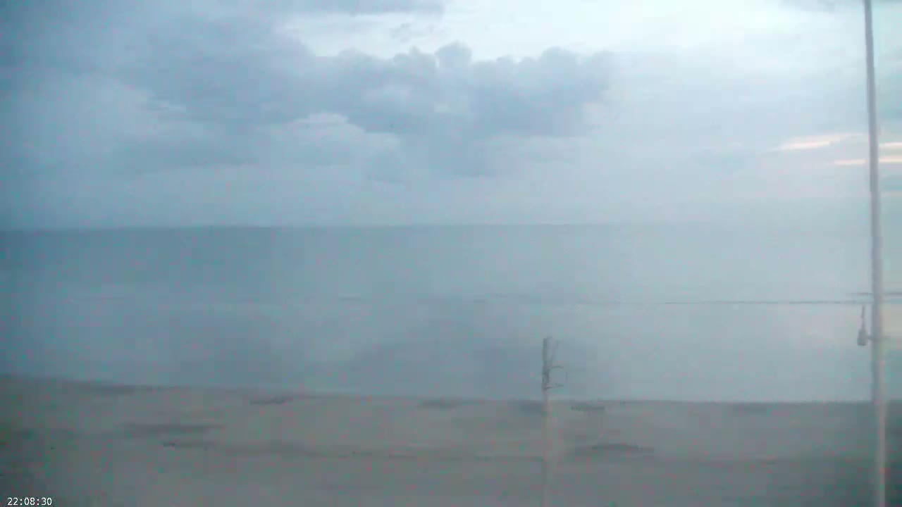 A hazy, overcast sky hangs over a calm, light-blue ocean meeting a flat, light-grey beach.