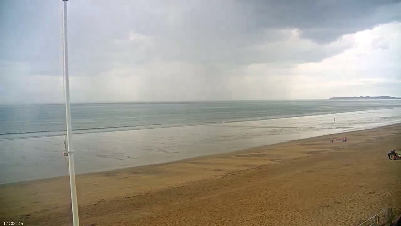 A wide sandy beach stretches to a calm sea under a cloudy, overcast sky with light rain.