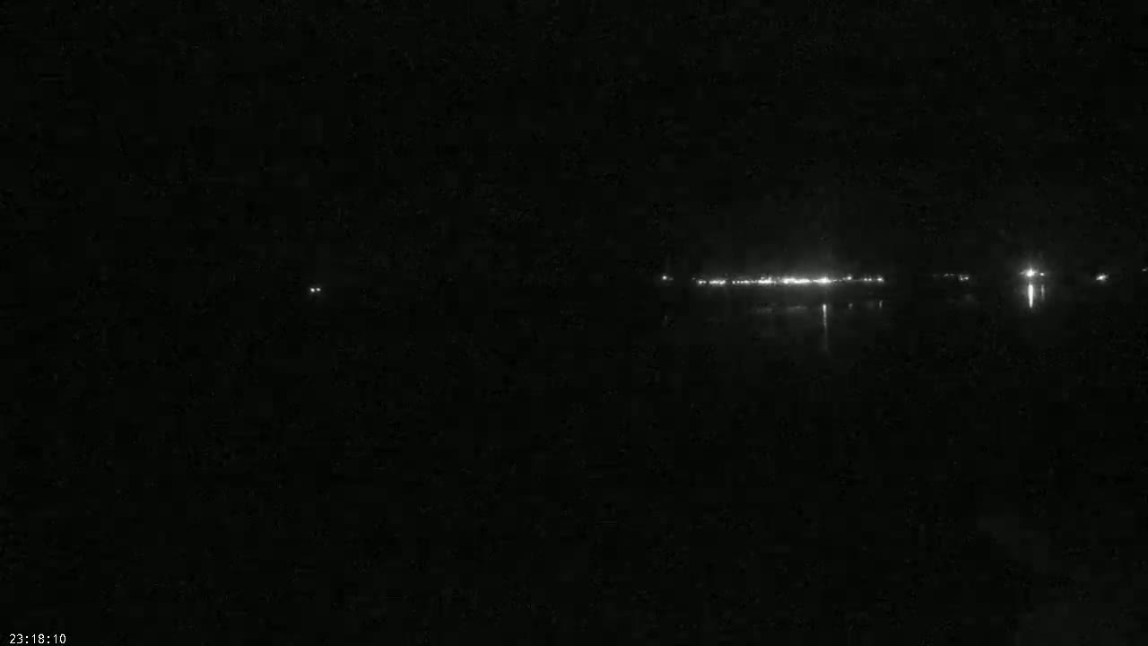A dark, nighttime scene shows a distant line of lights reflected on a calm body of water.
