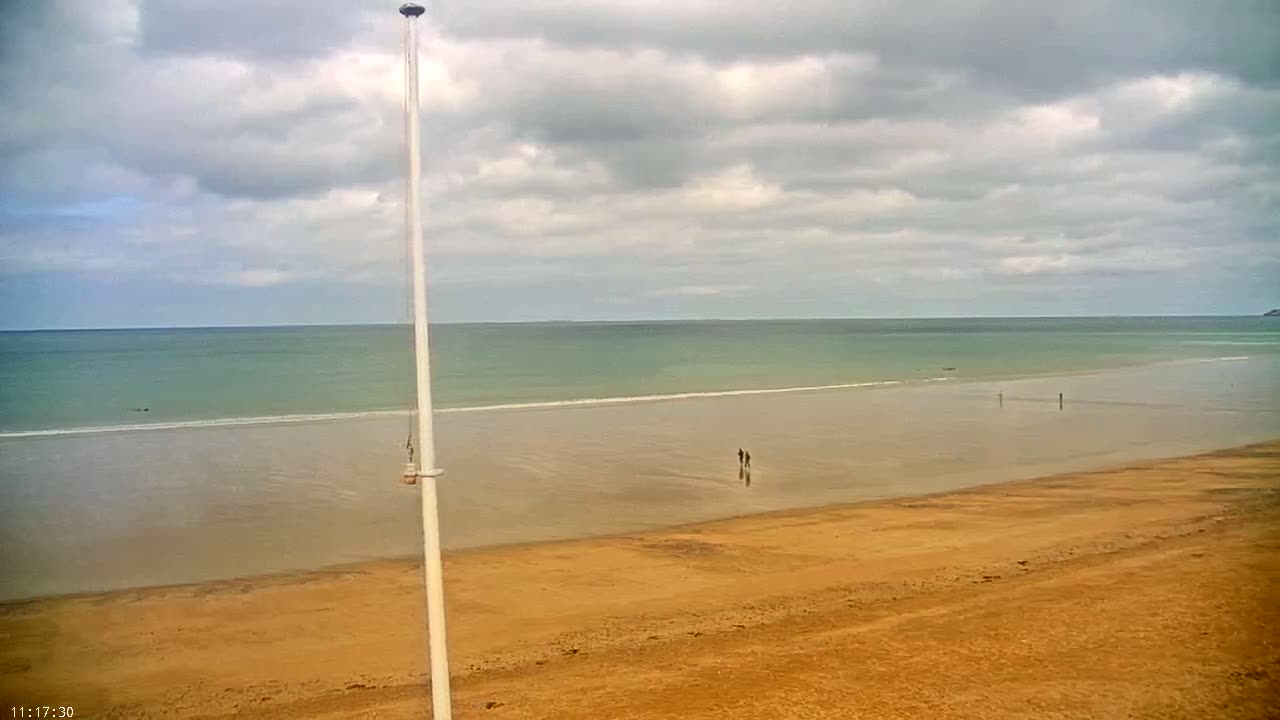 A tan, expansive beach stretches to a calm, teal ocean under a cloudy sky; a tall white pole stands in the foreground.