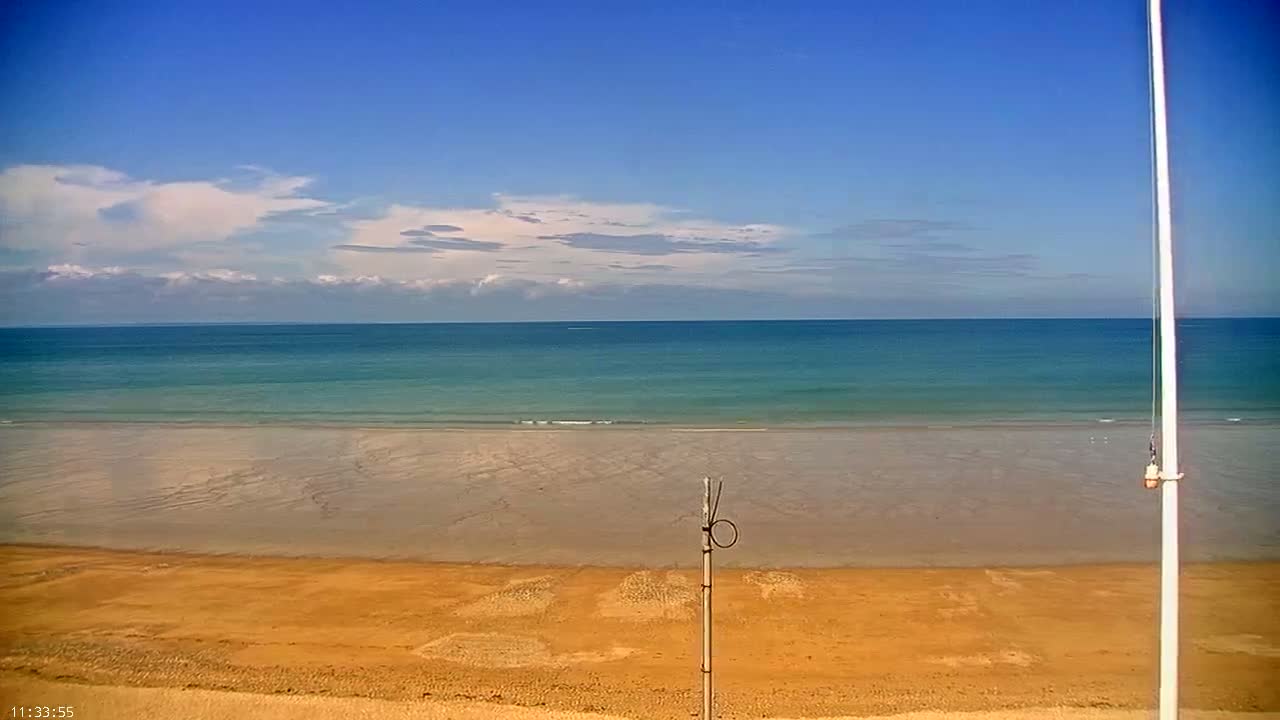 A sandy beach stretches to a calm, teal ocean under a mostly sunny sky with scattered clouds.
