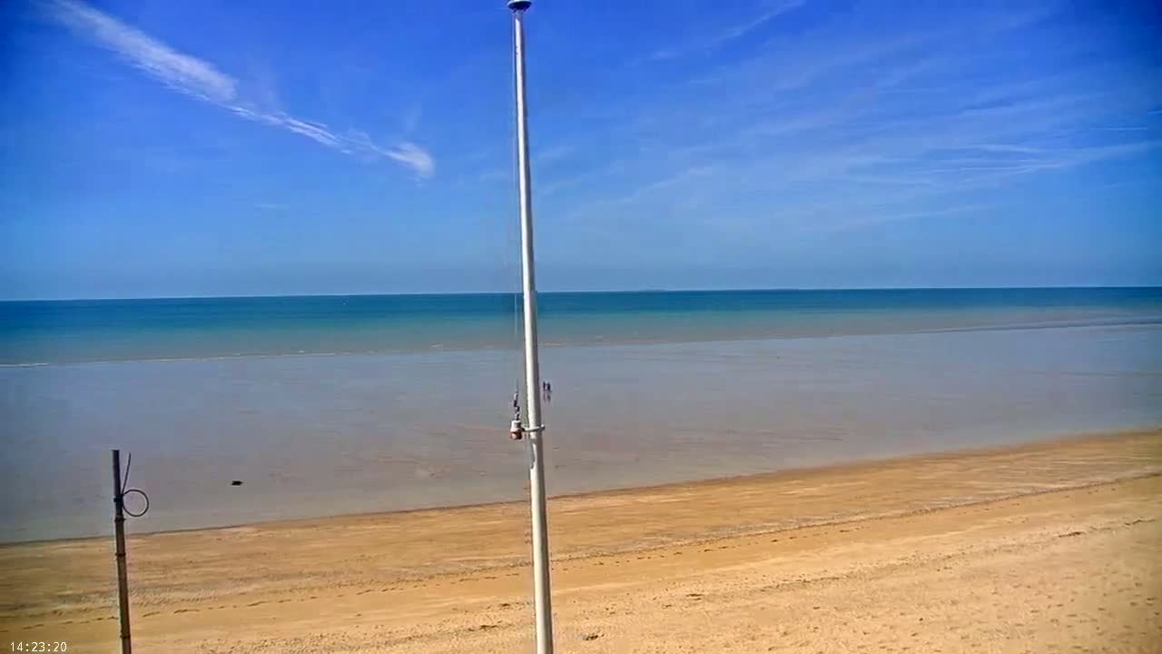 A tall flagpole stands on a sandy beach overlooking a calm, blue ocean under a clear, mostly sunny sky.