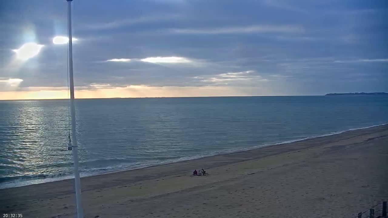 A sandy beach slopes down to a calm sea under a partly cloudy sky, with sun rays breaking through the clouds and a small group of people sitting on the sand.