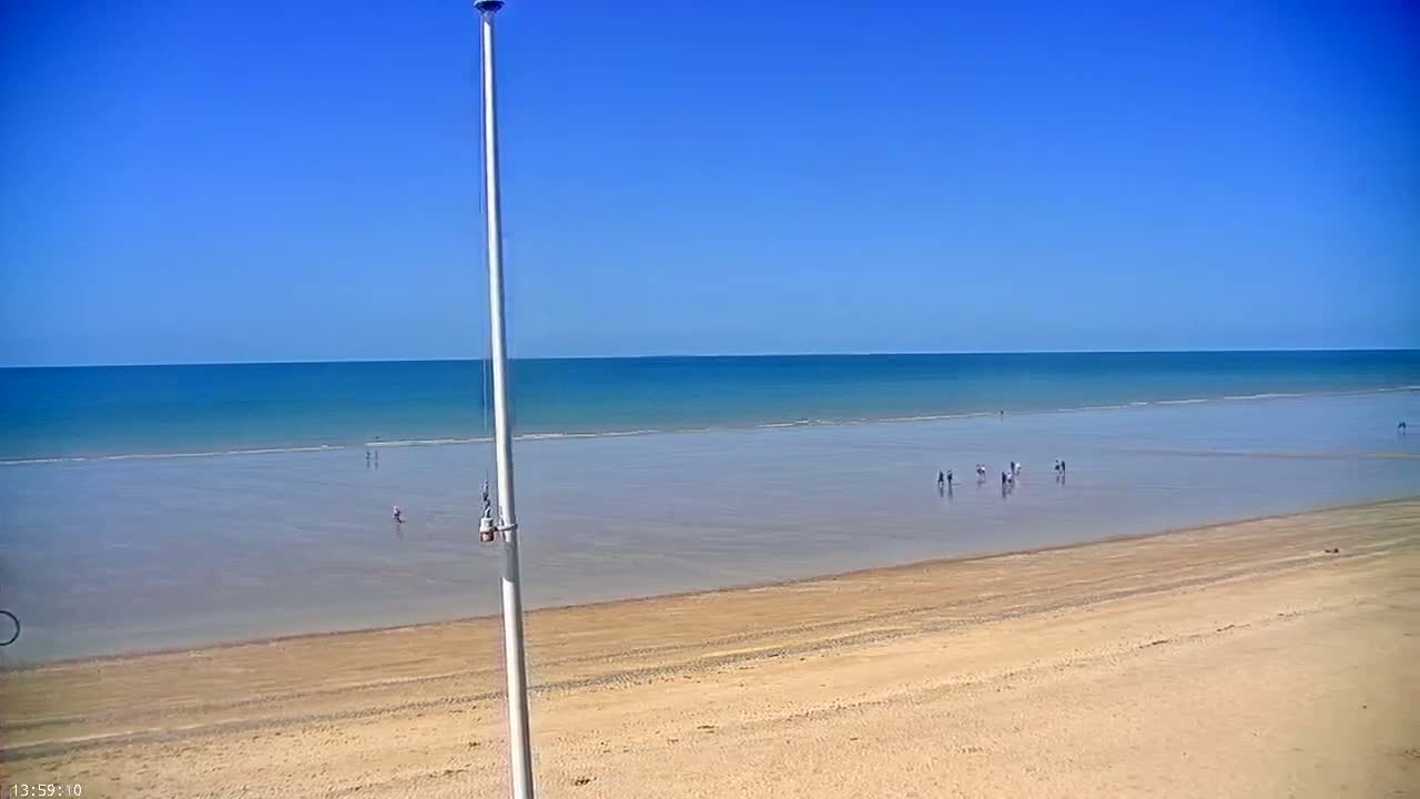 A wide sandy beach stretches to a calm, blue ocean under a clear blue sky, with a few people visible on the sand and in the shallow water.