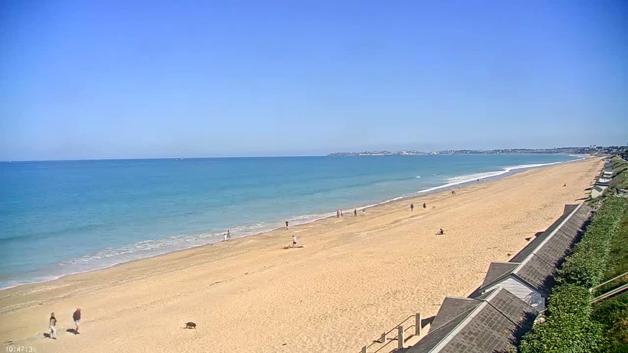 A long sandy beach stretches to a calm, blue ocean under a clear blue sky, with a few people scattered along the shore and several beach huts lined up on the right.