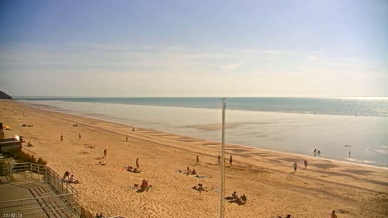 A wide sandy beach with scattered people stretches to a calm, light-blue ocean under a clear, sunny sky.