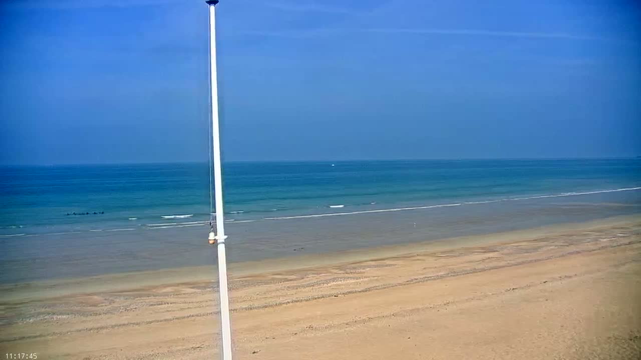 A tall white pole stands in the foreground of a sandy beach under a clear blue sky, with calm ocean waves gently lapping the shore.