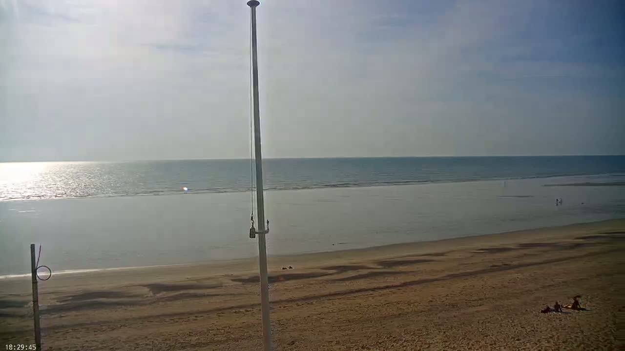 A sandy beach stretches to a calm, sunlit ocean under a mostly clear sky, with a tall pole standing in the foreground.