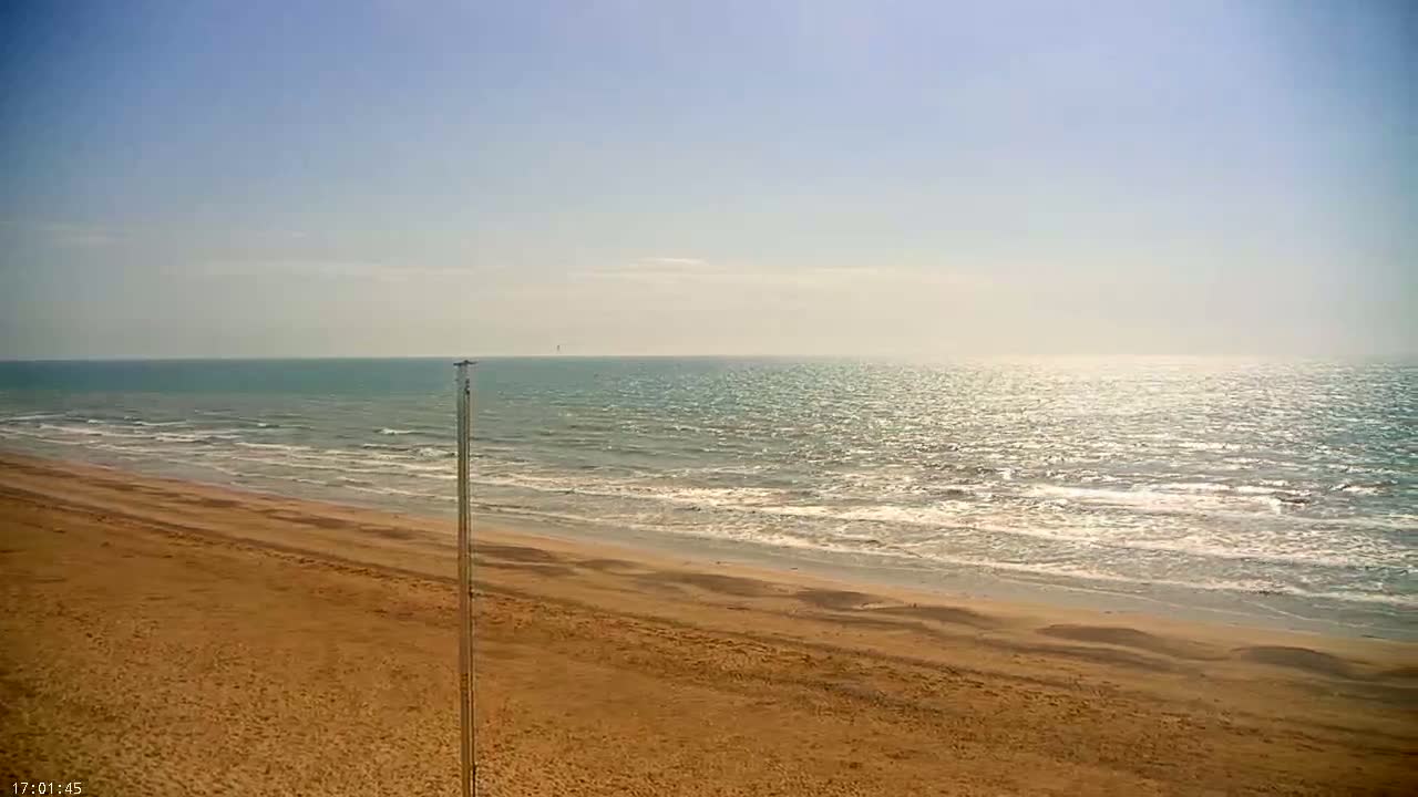 A wide sandy beach stretches to a calm, sunlit ocean under a clear, light blue sky.