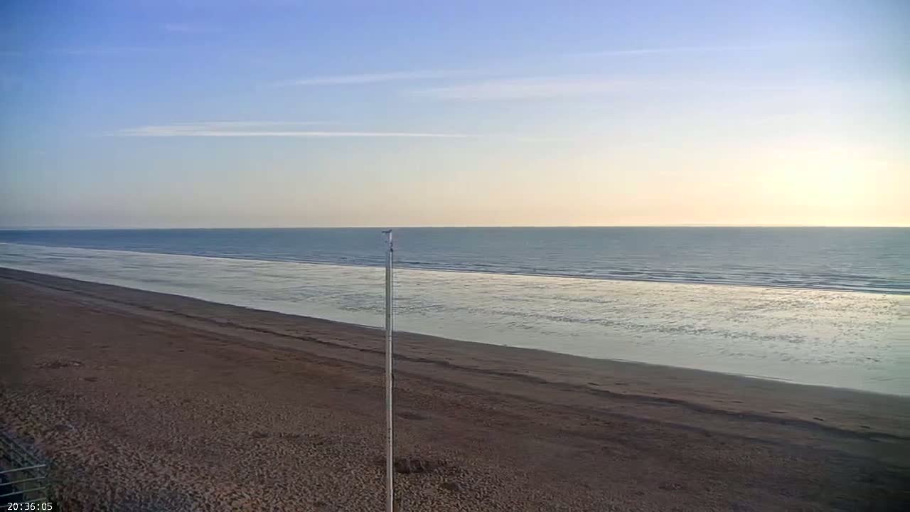 A long, sandy beach stretches towards a calm ocean under a clear, pale blue sky.