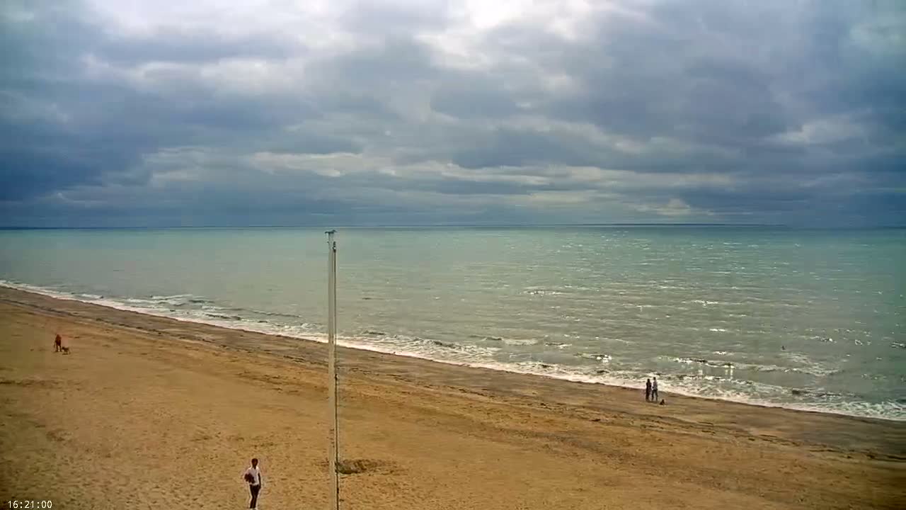 A sandy beach slopes down to a calm, light-green sea under a cloudy sky.