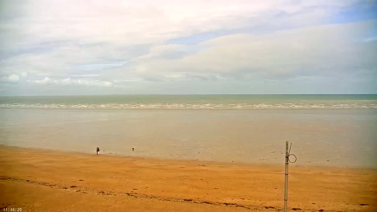 A person walks on a wide, orange-tan sandy beach toward a calm, light-green ocean under a mostly cloudy sky.