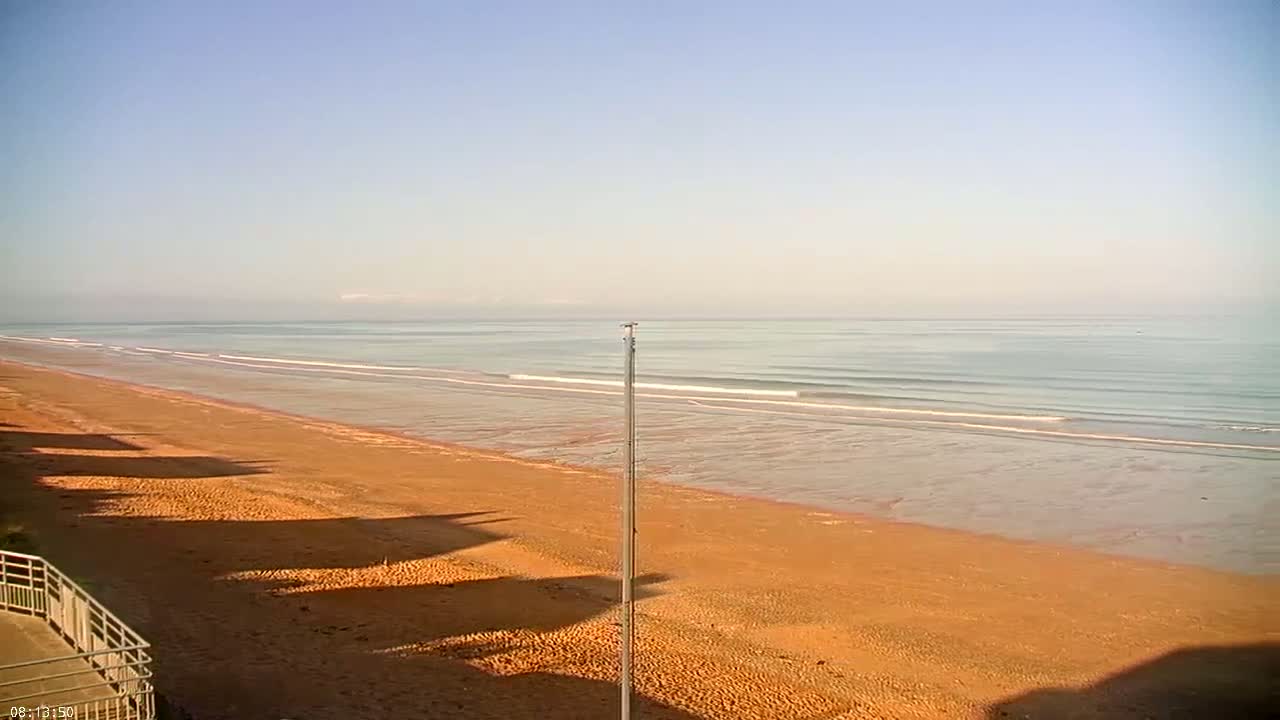 A long, orange-sand beach stretches towards a calm, light-blue ocean under a clear, pale-blue sky.