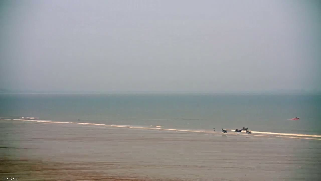 A hazy, overcast day shows a wide, flat expanse of light brown sand and shallow, calm water leading to a calm sea.