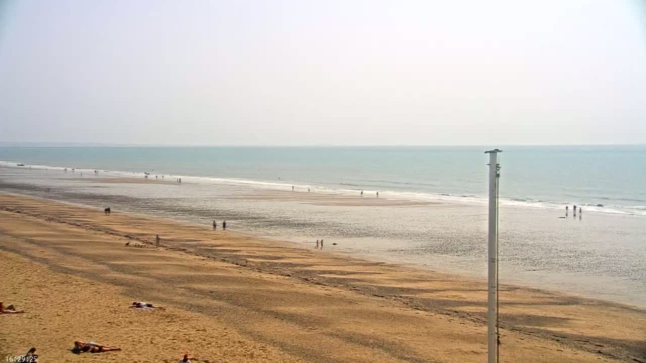 A long, sandy beach stretches to a calm, light-blue ocean under a hazy sky, with a few people scattered across the sand and in the water.