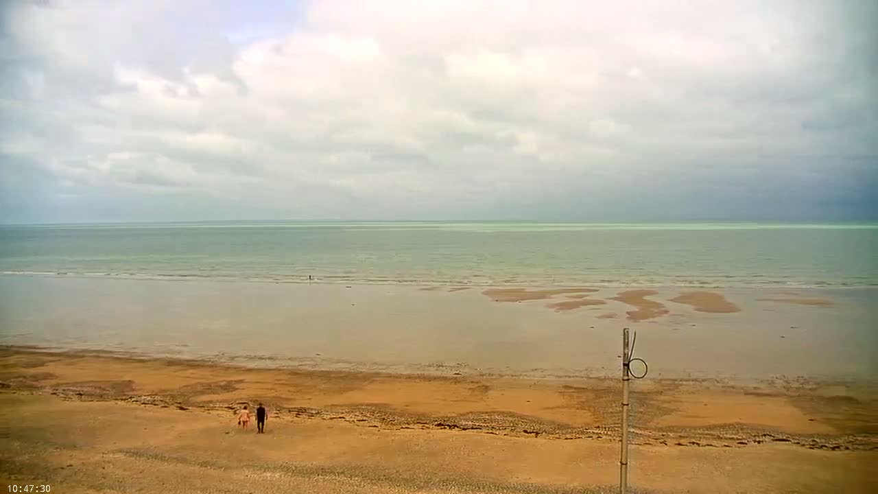 A wide, sandy beach stretches to a calm, pale green sea under a mostly cloudy sky.