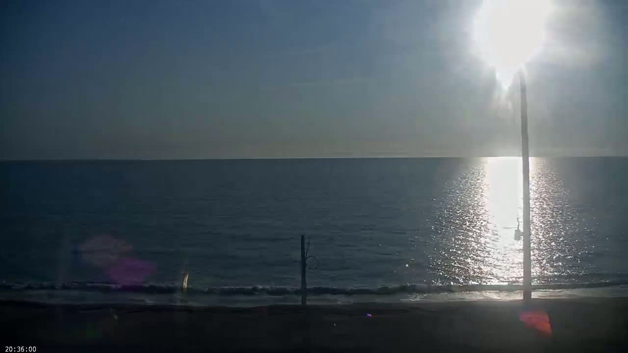 A bright sun shines over a calm ocean meeting a dark sandy beach with a tall, thin post standing near the water's edge.