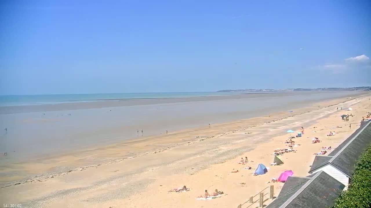 A wide sandy beach stretches to a calm ocean under a clear blue sky, with people relaxing and swimming on a sunny day.