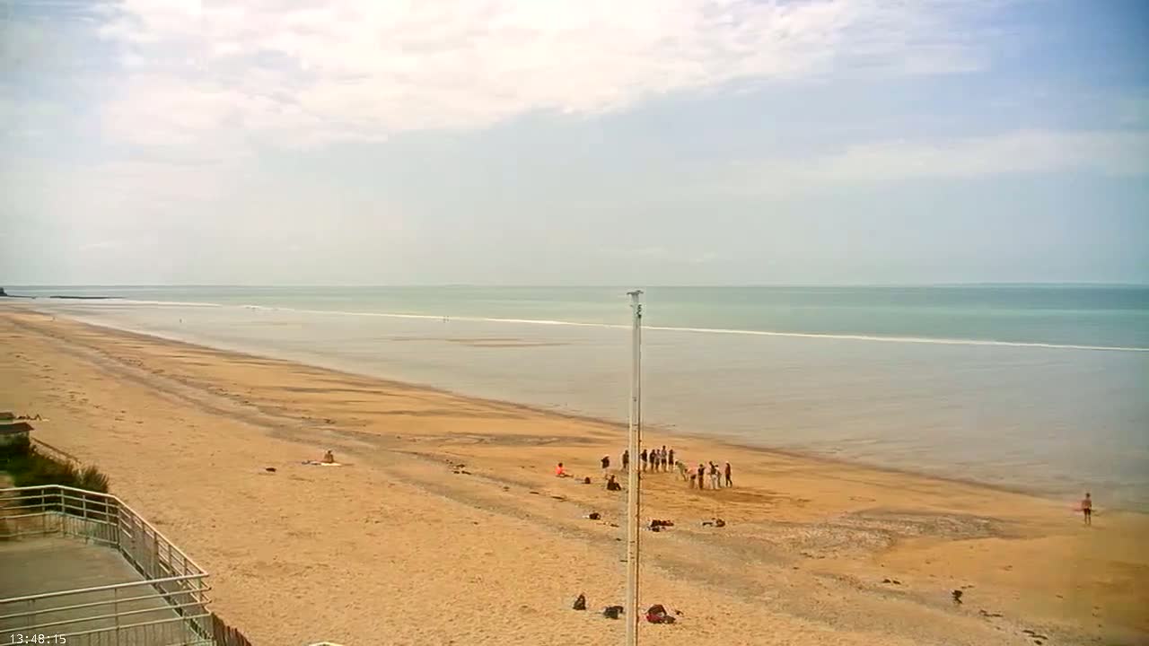 A long, sandy beach stretches to a calm, light-green ocean under a mostly cloudy sky, with a small group of people gathered near the water's edge.