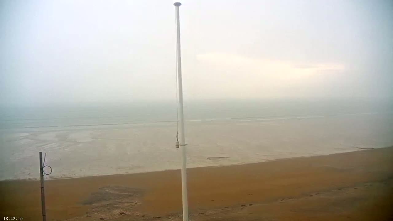 A tall flagpole stands on a sandy beach overlooking a calm, foggy sea.